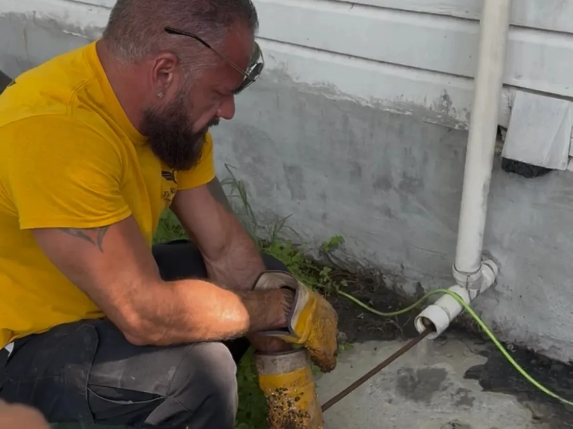 Tito Pacheco metiendo un cable de destapar por un registro de drenaje afuera de una casa para destapar la línea principal